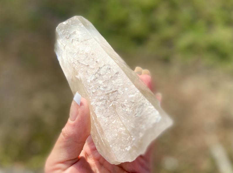 May include: A clear quartz crystal point with a textured surface. The crystal is held in a hand, with a blurred green background. The crystal is a light beige color and has a natural, raw appearance. The crystal is a natural mineral.