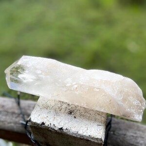 May include: A clear quartz crystal with a natural, rough texture. The crystal is a light, translucent color and rests on a weathered, white-painted wooden block. The background is a blurred green.