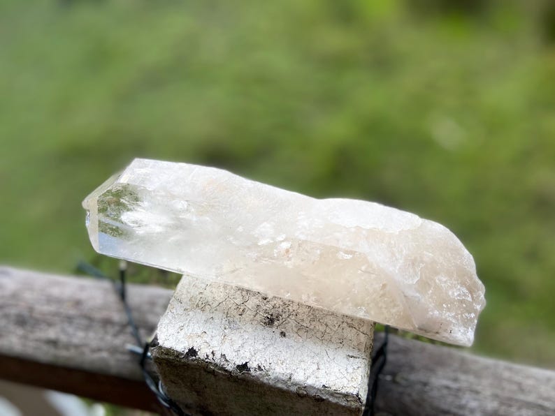 May include: A clear quartz crystal with a natural, rough texture. The crystal is a light, translucent color and rests on a weathered, white-painted wooden block. The background is a blurred green.