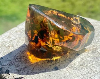 May include: A polished, translucent amber ring featuring deep golden and honey-colored hues with visible organic inclusions. The ring has a smooth, rounded surface that catches the light, showcasing a warm, multi-tonal appearance throughout the resin.