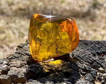 May include: A polished, translucent amber gemstone with a warm, golden-yellow hue. The stone features a smooth, multifaceted surface that catches the light, revealing internal organic inclusions and tiny air bubbles trapped within the resin.