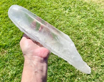 May include: A large, elongated, clear quartz crystal point with a smooth, translucent surface and natural facets. The crystal exhibits internal inclusions and a tapered shape, showcasing a glassy texture and a colorless, transparent appearance.