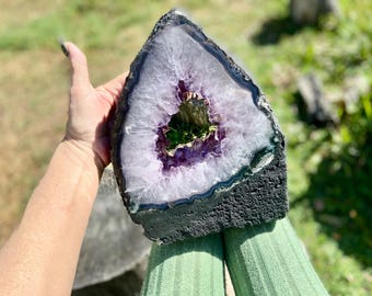 May include: A large, amethyst geode with a dark, textured exterior. The interior features a crystalline structure with shades of purple and white. The geode is held in a hand against a green background.