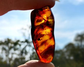 May include: A translucent amber gemstone, held between fingers, showcasing a blend of deep red and golden yellow hues. The polished surface reveals internal patterns and inclusions, with light passing through, creating a glowing effect. The background is a blurred view of a blue sky and trees.