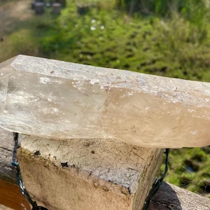 May include: A large, clear quartz crystal with a pointed tip. The crystal is a light, translucent color and rests on a weathered, light-colored brick. The background features green grass and sunlight.