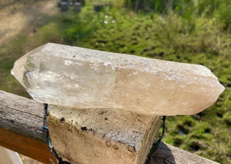 May include: A large, clear quartz crystal with a pointed tip. The crystal is a light, translucent color and rests on a weathered, light-colored brick. The background features green grass and sunlight.