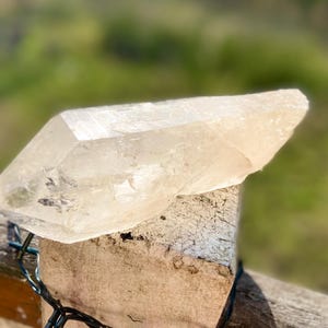 May include: A large, clear quartz crystal with a pointed tip rests on a weathered wooden post. The crystal is translucent, allowing light to pass through, and has a natural, rough texture. The background is blurred, suggesting an outdoor setting.