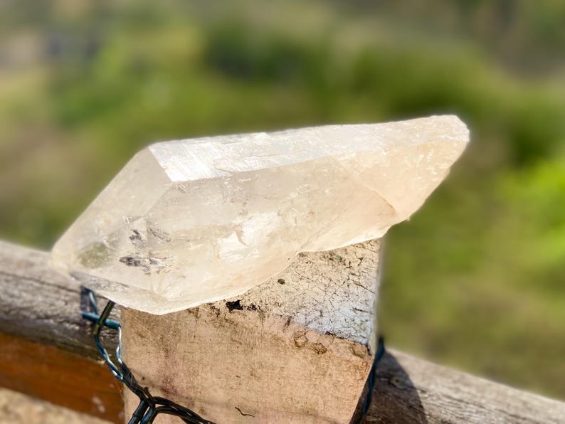 May include: A large, clear quartz crystal with a pointed tip rests on a weathered wooden post. The crystal is translucent, allowing light to pass through, and has a natural, rough texture. The background is blurred, suggesting an outdoor setting.