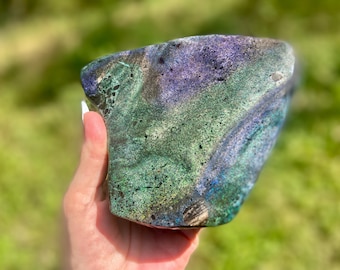 May include: A large, irregular mineral specimen featuring a vibrant blend of deep purple, forest green, and teal hues. The surface displays a textured, crystalline appearance with swirling color patterns and small dark inclusions throughout the stone.
