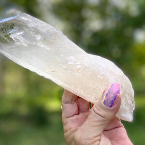 May include: A large, clear quartz crystal with a rough, natural texture. The crystal is held against a blurred green background, showcasing its translucent quality and natural form. The crystal is a light, neutral color.