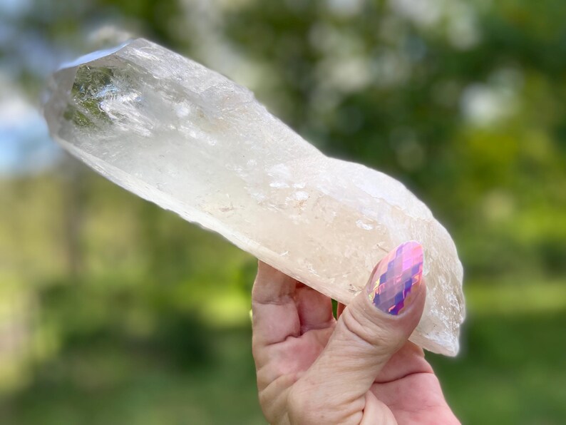 May include: A large, clear quartz crystal with a rough, natural texture. The crystal is held against a blurred green background, showcasing its translucent quality and natural form. The crystal is a light, neutral color.