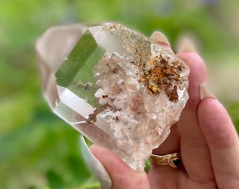 May include: A clear quartz crystal with a faceted tip and rough, textured base. The crystal has inclusions and a light brown and gold coloration on its surface. The background is a soft green.
