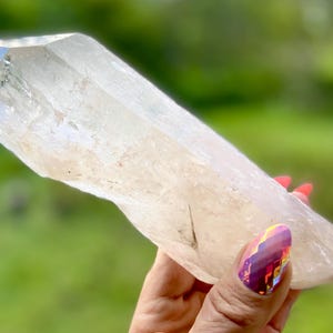 May include: A clear quartz crystal held in a hand with red nail polish. The crystal is elongated with a pointed tip and a rough, natural texture. The background is a blurred green.