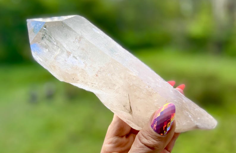 May include: A clear quartz crystal held in a hand with red nail polish. The crystal is elongated with a pointed tip and a rough, natural texture. The background is a blurred green.