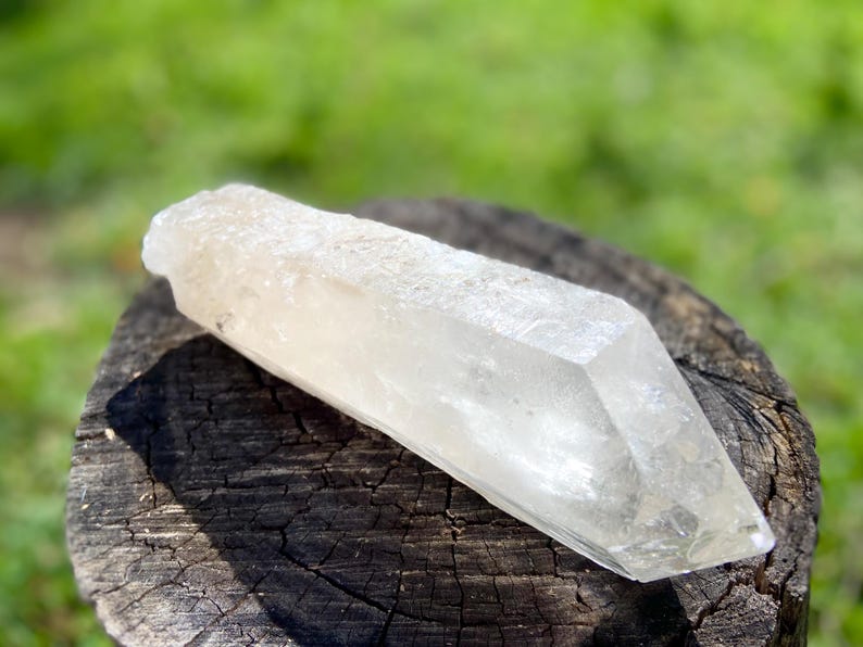May include: A clear quartz crystal point, resting on a weathered wooden surface. The crystal is translucent with a textured surface and a sharply defined point. The background is blurred green foliage, suggesting an outdoor setting.