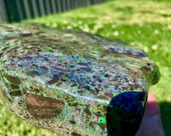 May include: A polished slab of rock featuring a complex mosaic of dark brown, black, and grey angular inclusions embedded within a vibrant matrix of shimmering green and blue iridescent flecks that catch the light across the smooth surface.