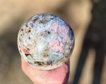 May include: A polished crystal sphere with a mosaic pattern of pink, blue, and cream colors. The sphere is held in a hand, showcasing its intricate design and natural beauty. This decorative sphere is a unique home decor item.