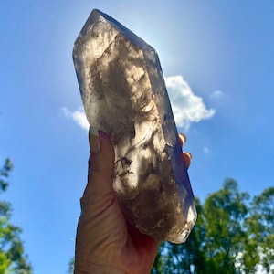 Puede incluir: Un gran cristal de cuarzo ahumado translúcido sostenido contra un cielo azul brillante con algunas nubes blancas. El cristal es de color marrón claro y tiene una punta afilada. El sol brilla a través del cristal, creando un efecto brillante.
