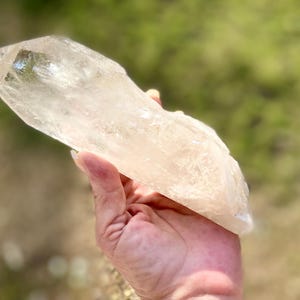 May include: A large, clear quartz crystal with a pointed tip and a rough, textured surface. The crystal is held in a hand against a blurred green and brown background. The crystal appears to be approximately 6 inches long.