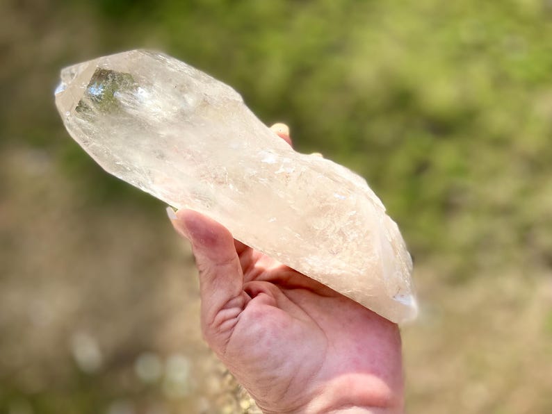 May include: A large, clear quartz crystal with a pointed tip and a rough, textured surface. The crystal is held in a hand against a blurred green and brown background. The crystal appears to be approximately 6 inches long.