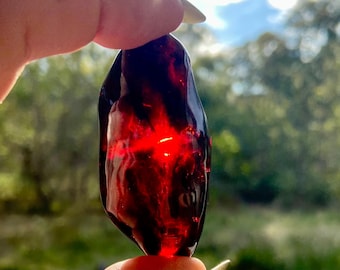 May include: A dark red, translucent gemstone with a polished surface. The stone is held between fingers, with a bright red light emanating from within. The background is blurred, showing green foliage and a blue sky.