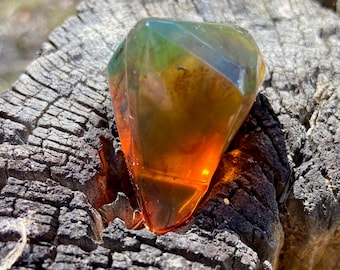 May include: A faceted, translucent gemstone with a gradient color transition from deep orange at the pointed base to a vibrant green at the top. The object has a geometric, diamond-like shape with multiple flat surfaces that catch the light.