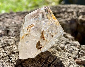 May include: A clear, multifaceted quartz crystal point featuring internal fractures and natural golden-brown mineral inclusions. The stone has a pointed, geometric shape with sharp edges and a translucent, glass-like appearance throughout its structure.