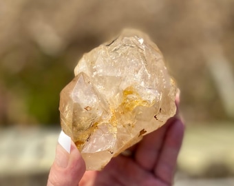 May include: A raw, natural quartz crystal with a light amber color. The crystal has a rough, textured surface and a few small inclusions. The quartz is held in a hand, with a blurred background.