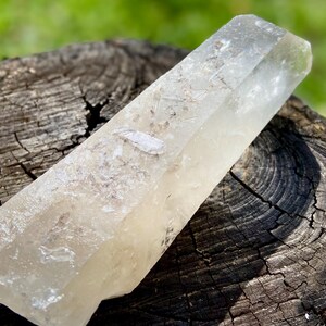 May include: A clear quartz crystal point, resting on a weathered wooden surface. The crystal is elongated with a slightly cloudy appearance and a natural, unpolished texture. The background is blurred green grass.