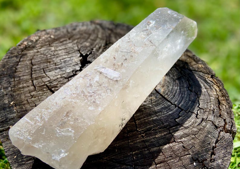 May include: A clear quartz crystal point, resting on a weathered wooden surface. The crystal is elongated with a slightly cloudy appearance and a natural, unpolished texture. The background is blurred green grass.