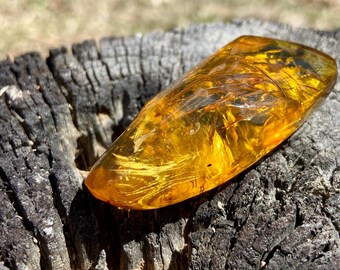 May include: A polished, translucent piece of golden-yellow amber with a smooth, elongated, and slightly irregular shape. The interior displays visible natural inclusions, fine fibrous patterns, and internal fractures that catch the light.