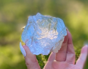 May include: A light blue, translucent crystal with a jagged, uneven form. The crystal is held in a hand, set against a blurred green backdrop, implying an outdoor environment. The crystal's surface catches the light, producing a sparkling appearance.