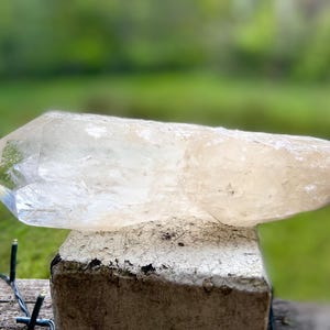 May include: A large, clear quartz crystal with a pointed tip, resting on a weathered, light-colored stone. The crystal has a slightly translucent appearance, with subtle color variations. The background is a blurred green, suggesting an outdoor setting.