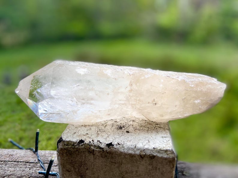 May include: A large, clear quartz crystal with a pointed tip, resting on a weathered, light-colored stone. The crystal has a slightly translucent appearance, with subtle color variations. The background is a blurred green, suggesting an outdoor setting.