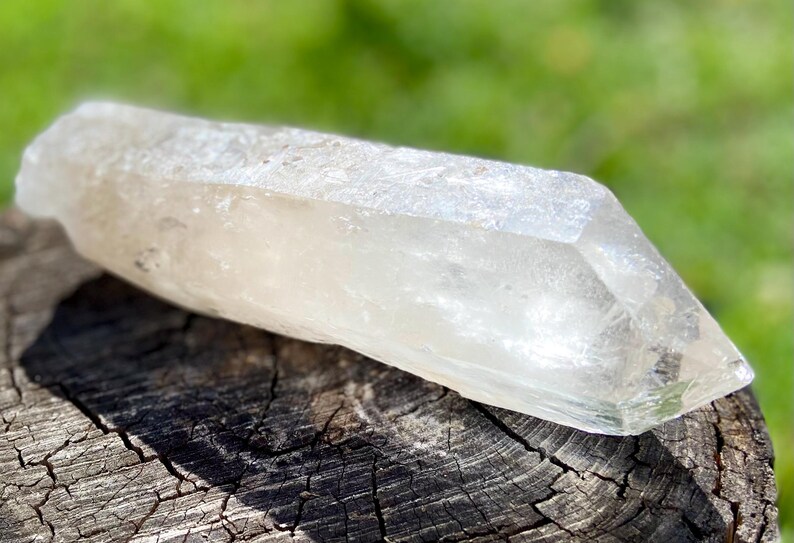 May include: A clear quartz crystal, elongated and faceted, rests on a weathered wooden surface. The crystal's translucent quality allows light to pass through, highlighting its natural form. The background is a soft green, suggesting an outdoor setting.