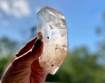 May include: A large, clear quartz crystal with a natural, raw shape. The crystal has a slight pink hue and is translucent. It is held in a hand against a blurred background of blue sky and green foliage.