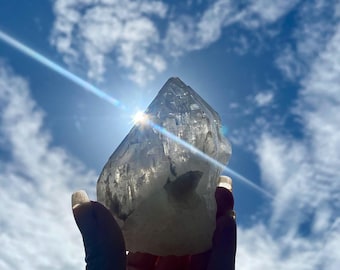 May include: A clear quartz crystal held up against a bright blue sky with fluffy white clouds. The sunlight shines through the crystal, creating a sparkling effect. The crystal is held in a hand.