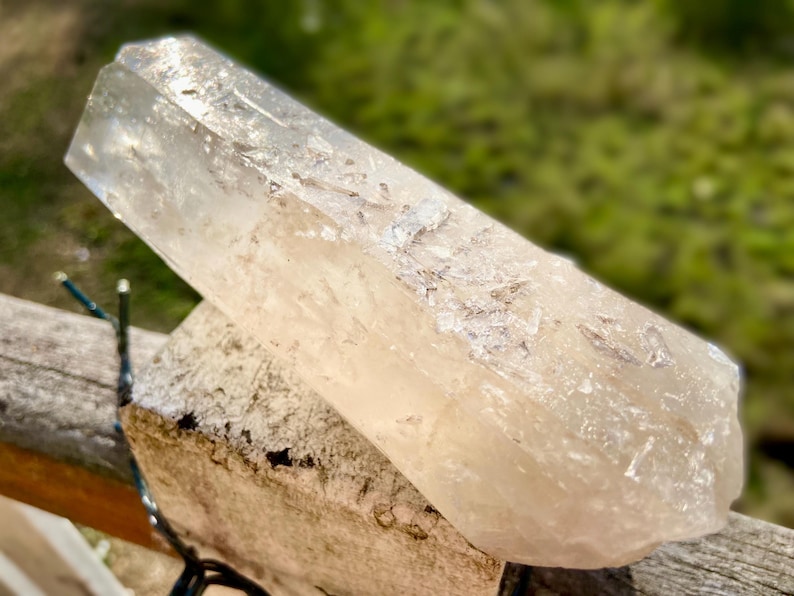 May include: A large, clear quartz crystal with a slightly milky appearance. The crystal is elongated and rests on a rough, light-colored stone. The background is blurred, with green foliage visible.