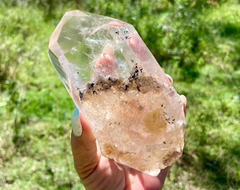 May include: A clear quartz crystal with a light pink hue and inclusions of black and brown minerals. The crystal is held up against a backdrop of green grass and sunlight.