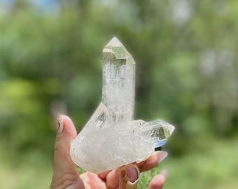 May include: A clear quartz crystal cluster with a large, pointed central crystal and two smaller crystals. The crystals are translucent with visible internal structures. The background is a blurred green, suggesting an outdoor setting.