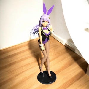May include: A figurine of a female anime character in a purple bunny suit with matching ears. The figure has long lavender hair, a black vest, and is standing on a black circular base. The character is winking and making a peace sign.