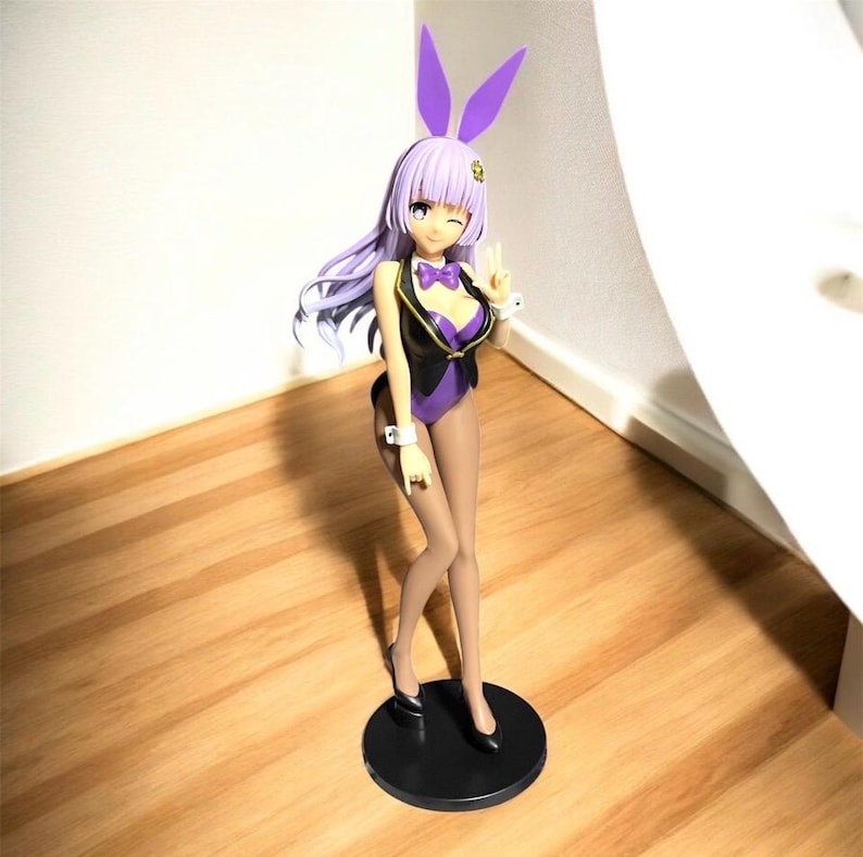 May include: A figurine of a female anime character in a purple bunny suit with matching ears. The figure has long lavender hair, a black vest, and is standing on a black circular base. The character is winking and making a peace sign.