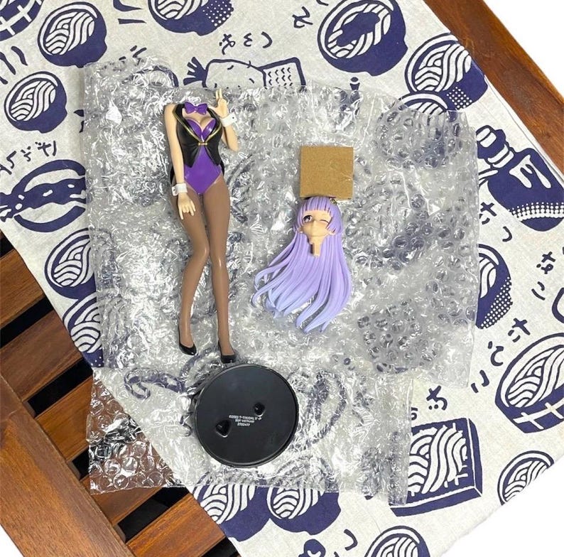 May include: A disassembled anime figure featuring a purple bodysuit, black vest, and long brown legs. The figure includes a separate head with long, light purple hair, a black base, and a small cardboard piece. The figure is packaged in bubble wrap.