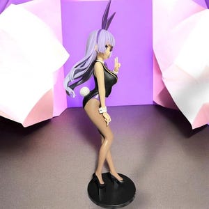 May include: A figurine of a woman in a black bunny suit with purple accents, including a matching headband and ears. The figure is posed on a black circular base, with a white fluffy tail and long, flowing lavender hair.