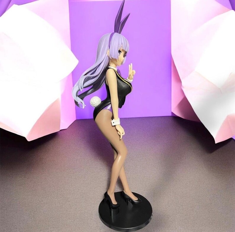 May include: A figurine of a woman in a black bunny suit with purple accents, including a matching headband and ears. The figure is posed on a black circular base, with a white fluffy tail and long, flowing lavender hair.