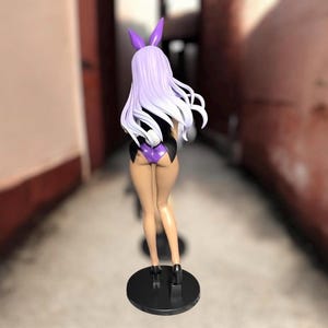 May include: A figurine of a woman in a bunny costume, viewed from the back. The figure has long, light purple hair and wears a black bodysuit with purple accents. The figurine stands on a black circular base.