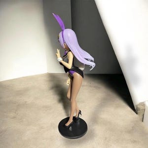 May include: A figurine of a woman in a bunny suit, featuring a black and purple outfit, high heels, and a purple bunny ear headband. The figure is standing on a black circular base, with long, flowing lavender hair. The figure is making a peace sign.