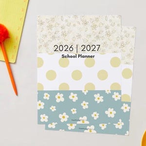 May include: A school planner for 2026 and 2027 with a floral and polka dot design. The planner has a light blue floral pattern at the bottom, a white polka dot pattern in the middle, and a cream floral pattern at the top. An orange pen and notepad are on the left.