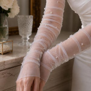 May include: Sheer white tulle opera-length gloves featuring a ruched design and scattered small white faux pearls throughout the fabric. The gloves extend past the elbows and are constructed from a lightweight, semi-transparent mesh material.