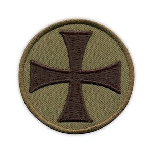 Puede incluir: Parche circular con un diseño de cruz marrón oscuro sobre un fondo verde oliva. La cruz es bordada. El parche está bordeado con costuras verde oliva más claras. El diseño recuerda a un emblema militar.